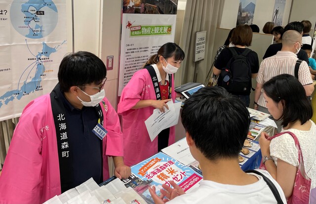 北海道森町と書かれたピンク色の法被を着た男女が、チラシなどが置かれたテーブルをはさんで来場者へ説明をしている展示の様子の写真