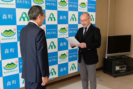 森町のバックボードの前に、眼鏡をかけたスーツ姿の男性2名が向かい合って立ち、右側の男性が手に持った紙に書かれた内容を読み上げている写真