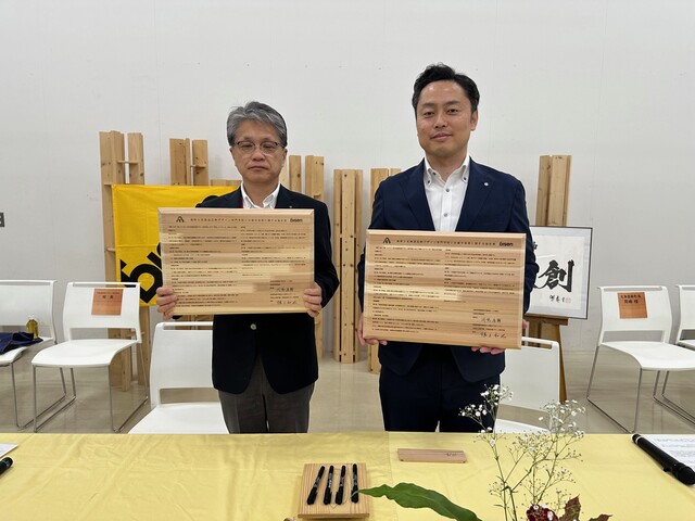 黄色いクロスがかかったテーブルの後ろに立った岡嶋町長と根上校長が、木の板でできた協定書をそれぞれ両手で持っている記念写真