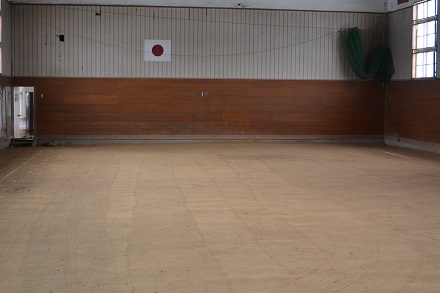 壁に日の丸が掲げられた天井の高い、土が敷かれた屋内土間練習場内の写真